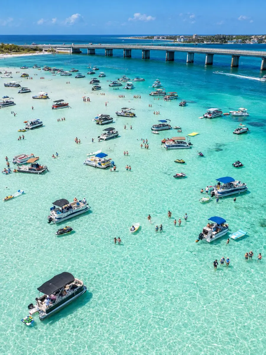 Aerial view of boats and jet skis anchored in turquoise shallows over a sandbar near a low bridge