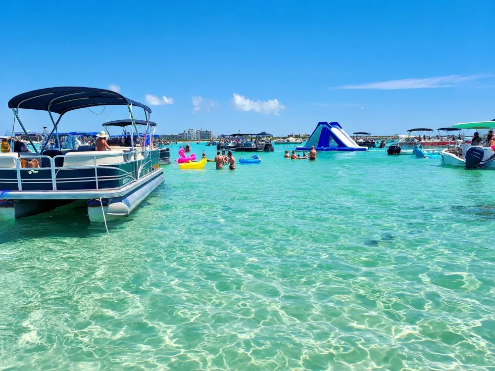 Boats anchored in knee-deep turquoise water over a sandbar, with floats and a waterslide