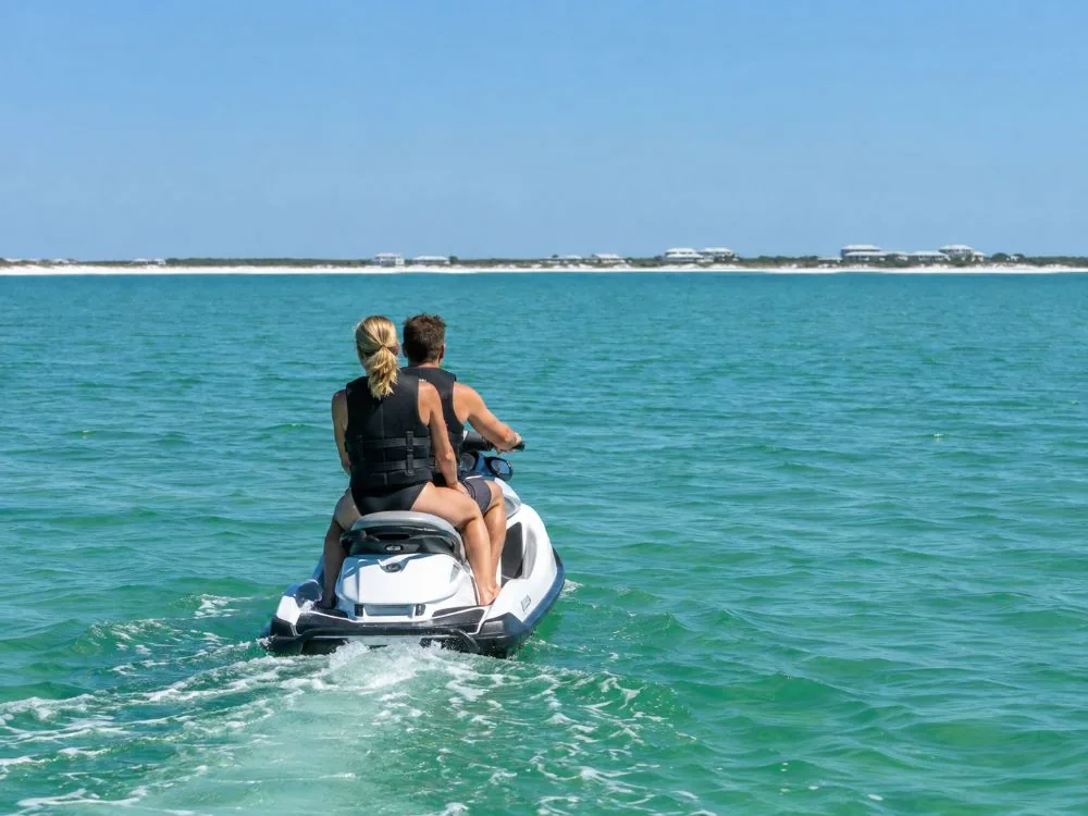Two adults in life jackets riding one jet ski slowly across calm emerald water, seen from behind